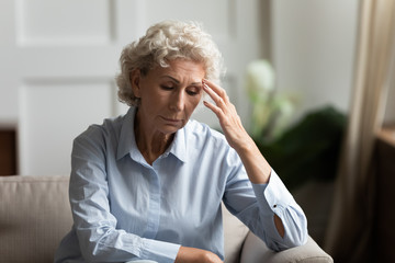 Stressed worried mature woman sit on sofa in living room lost in thoughts pondering or thinking,...