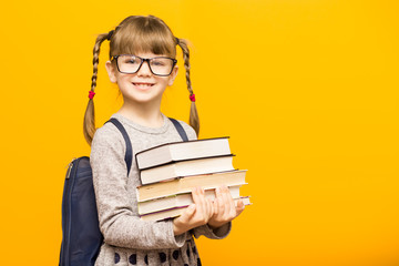 Back to school concept. Portrait of little smiling clever child girl in glasses is going to school for the first time with school backpack holding education books in hands isolated on yellow