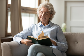 Happy mature woman sit relax on couch at home reading interesting paper book, smiling senior 60s...