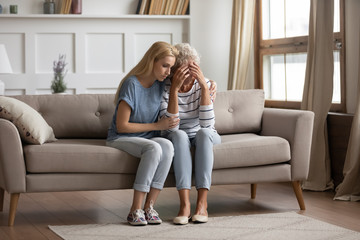 Loving grownup daughter sit on couch in living room hug support upset depressed senior mother,...