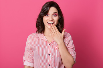 Indoor shot of surprised woman covering her mouth with hands while posing over pink background and looking at camera with opened mouth, female wearing casual outfit. People emotions concept.