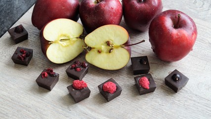 red ripe apples in drops of water and handmade dark chocolate candies decorated with dry berries on a wooden table on a black background
