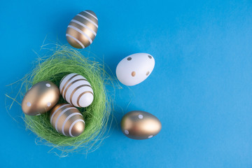 Easter eggs painted in gold colors in different patterns lie in a nest of green grass and next to it on a blue background. Easter holiday concept. Design for greeting card.