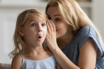 Young mother whisper in surprised cute little daughter ear tell secret, millennial mom or nanny play with small preschooler girl child, share close intimate moment at home, gossip or chat together