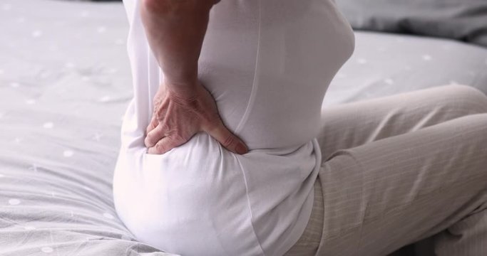 Senior grandma massaging back muscles sitting on bed. Old woman touching spine feeling backache pain suffering from morning discomfort in uncomfortable bed. Elder person arthritis concept. Close up