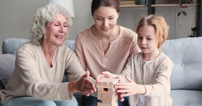 Multigenerational female family playing jenga board game together. Happy 3 three generations of women - old senior granny, young mother and small child granddaughter enjoying leisure activity at home.