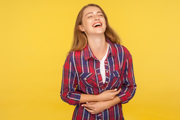 Portrait of carefree ginger girl in checkered shirt holding belly and laughing out loud after hearing hilarious joke, funny anecdote, being in good mood. studio shot isolated on yellow background