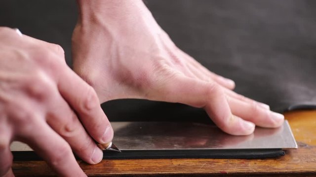 A tanner is cutting black leather sheet with a razor and metre. The master is making a leather belt for men.
