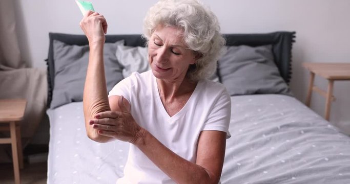 Smiling older woman applying moisturizer cream on elbow sitting on bed in the morning. Senior lady puts lotion doing everyday body dry skin hydration routine in bedroom. Elderly women skincare concept