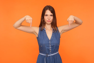 Must do here and right now. Angry brunette woman in denim dress pointing down with both hands, trying to control, demanding to come immediately. indoor studio shot isolated on orange background