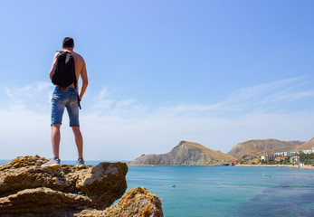 A sexy young man with a black backpack and a athletic body ,stands on a rock and looks at the blue water in the bay of the black sea, the back view. summer travel in vacation.