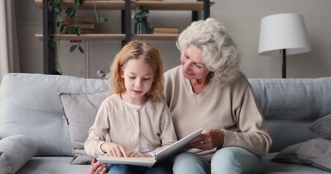Smart kid girl granddaughter learning reading with old grandma at home. Senior grandmother baby sitter teaching cute small grandkid of 6-7 years enjoying preschool elementary education hobby concept.