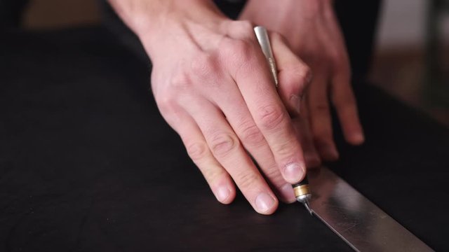 A tanner is cutting black leather sheet with a razor and metre. The master is making a leather belt for men.