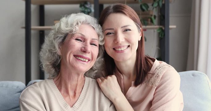 Relaxed happy young adult daughter embracing old mother and laughing together. Smiling grown granddaughter hugging senior grandma looking at camera at home. Two 2 age generations women family portrait