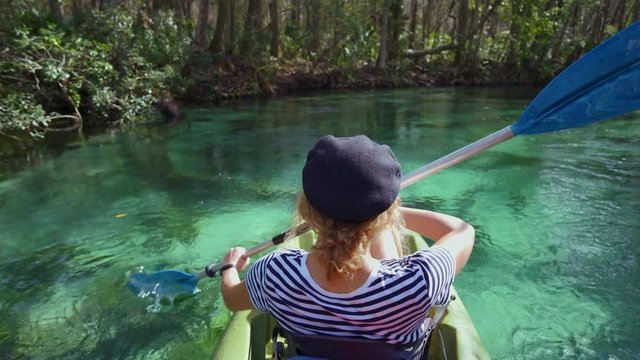 A young brunette model woman is paddling a kayak through the magical clear blue water of Weeki Wachee State Park Spring River. The sun is shining through green jungle trees during vacation.