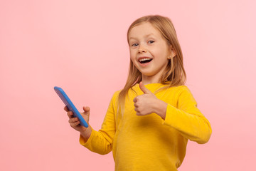 Best app for kids. Happy excited little girl holding smartphone and showing thumb up, like gesture...
