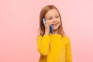 Cute little ginger girl with freckles smiling joyfully while calling parents on cell phone, good cellular, roaming, comfortable to use children mobile device. studio shot isolated on pink background