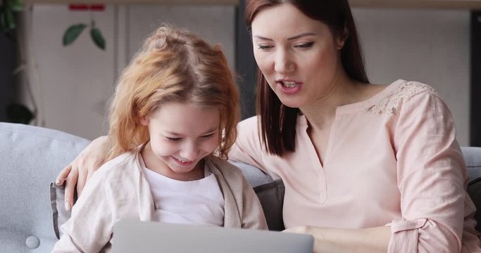 Happy child girl of 6-7 years learning using laptop computer modern tech with parental control. Young mom teaching kid daughter having fun surfing internet on wireless device sitting on sofa at home.