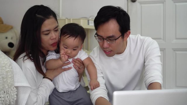 Asian father working using laptop with his wife and cute Asian baby boy while sitting on floor in cozy room at home.