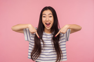 Wow look at advertise! Portrait of excited, surprised girl in striped t-shirt pointing down and looking with shocked face, showing place for commercial. indoor studio shoot isolated on pink background