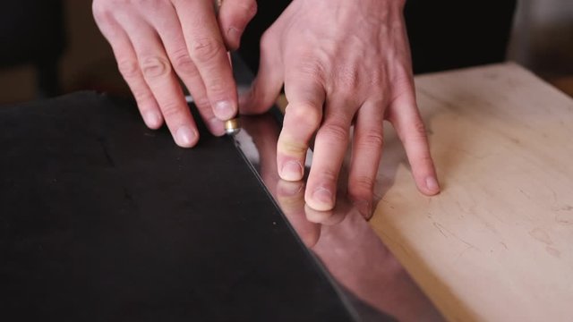 A tanner is cutting black leather sheet with a razor and metre. The master is making a leather belt for men.