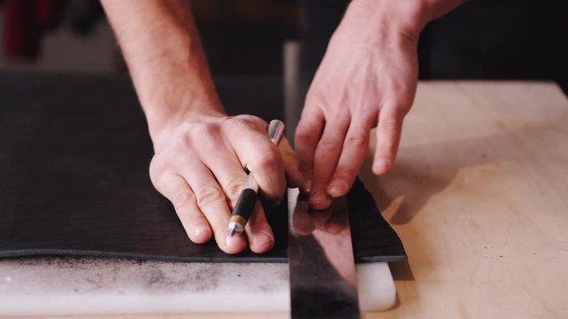 A tanner is cutting black leather sheet with a razor and metre. The master is making a leather belt for men.