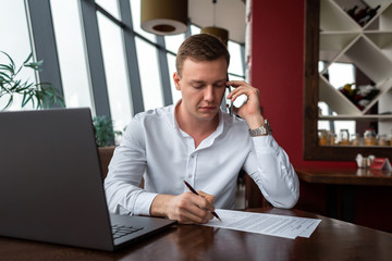 Plakat Young businessman wearing a white fashion shirt chatting and talking business on a mobile cell phone in a cafe with a laptop and sign documents. Freelance and selfemployment concept. Distance job.