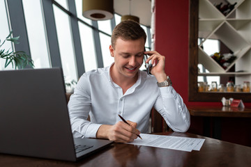Fototapeta na wymiar Young businessman wearing a white fashion shirt chatting and talking business on a mobile cell phone in a cafe with a laptop and sign documents. Freelance and selfemployment concept. Distance job.