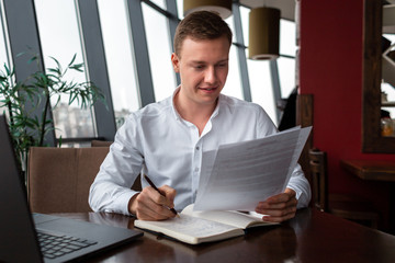 Fototapeta na wymiar Young businessman wearing a white fashion shirt sittting in a cafe with a laptop, documents and writing in notebook. Freelance and selfemployment concept. Distance job.