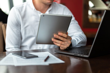 Closeup of hands of young businessman wearing a white fashion shirt with a tablet in a cafe with a laptop, phone and documents. Freelance and selfemployment concept. Distance job.