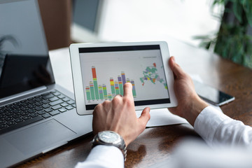 Closeup of hands with a tablet with diagrams of young businessman wearing a white fashion shirt in a cafe with a laptop and documents. Choose and pick options. Freelance and selfemployment concept.