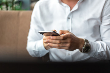 Closeup of hands of young businessman wearing a white fashion shirt with a mobile phone in a cafe with a laptop and documents. Freelance and selfemployment concept. Distance job.