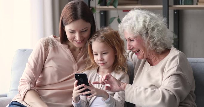 School kid girl daughter granddaughter mobile tech user holding smartphone showing funny social media video app to old grandmother and young mother. Three generation women family using gadget concept.