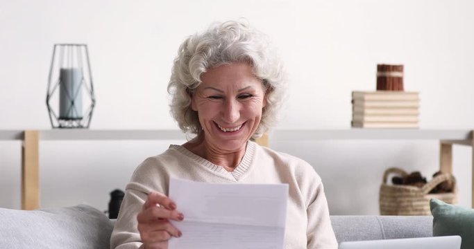 Cheerful old senior woman reads postal mail approval letter excited by good news. Excited elder lady feels satisfied with bill or tax refund, bank service offer, holding paper sitting on sofa at home