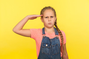 Yes sir! Portrait of responsible obedient little girl with braid in denim overalls saluting and looking at camera with respect, patriotic child. indoor studio shot isolated on yellow background