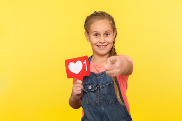 Hey you, like and comment child blog! Portrait of happy little girl in denim overalls holding social network heart button, follower notification symbol, popular internet content. studio shot isolated