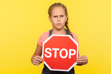 Forbidden way, prohibited to go. Portrait of angry little girl in denim overalls holding Stop symbol, showing red traffic sign and looking with dissatisfied expression. indoor studio shot isolated