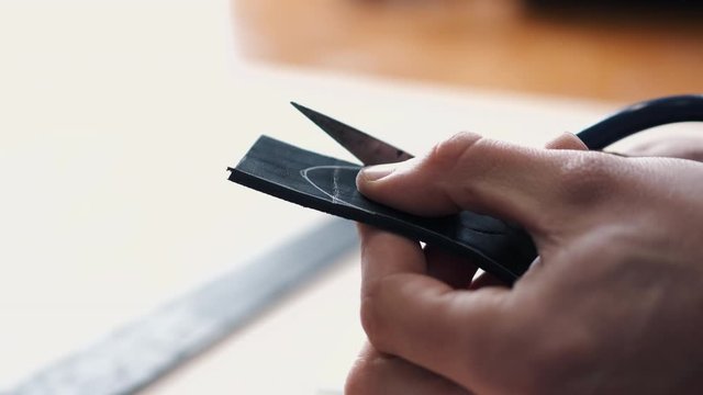 A leather craftsman is marking the end of a belt tail. The tanner is cutting the tail with scissors.