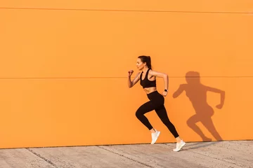 Zelfklevend Fotobehang Motivated confident fit woman athlete in tight sportswear, black pants and top, starting to run, jogging outdoor against orange wall, advertising area. Health care and weight loss, sport activity © khosrork