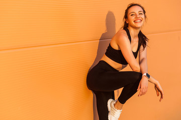 Attractive sexy girl with fit body in tight sportswear, black pants and top, leaning against orange wall and smiling to camera, living healthy sport life, full of energy. isolated on advertising area
