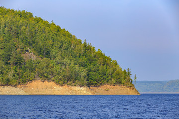 Obraz premium Beautiful landscape. Zeya reservoir, Amur region. A beautiful rocky shore with tall green trees goes into the calm blue waters of the reservoir.