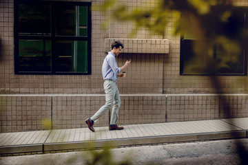Happy Young Businessman in Casual wear Using or reading Mobile Phone while Walking by the Urban Building. Lifestyle of Modern People. Side View. Full Length