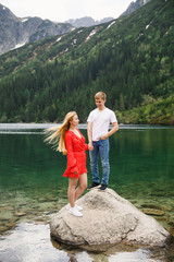 a young couple, a girl in a red dress, a man in a white T-shirt and blue pants stand on a background of mountains near the lake in the park