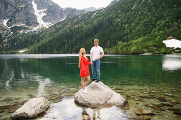 a young couple, a girl in a red dress, a man in a white T-shirt and blue pants stand on a background of mountains near the lake in the park