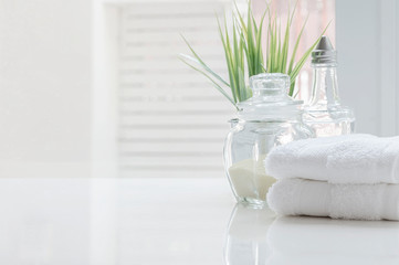 White folded towels and glass bottle on white table with copy space on blurred bathroom background.