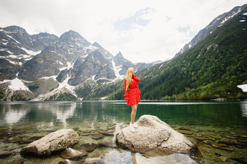 young girl in a red dress stands on a stone against the background of a lake and mountains in the Tatra National Park in Poland