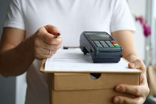 Close-up view of man holding cardboard box and payment terminal in hands. Credit card contactless paying for service. Package delivery and modern technology concept