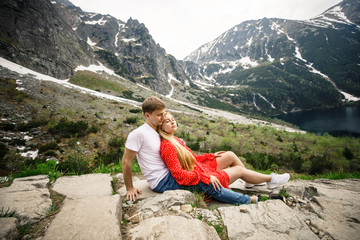 young beautiful couple, a girl in a red dress, and a man in a white T-shirt and blue pants, sitting on a stone on a background of mountains