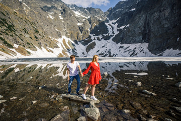 a young couple, a girl in a red dress, a man in a white T-shirt and blue pants stand on a background of mountains near the lake in the park