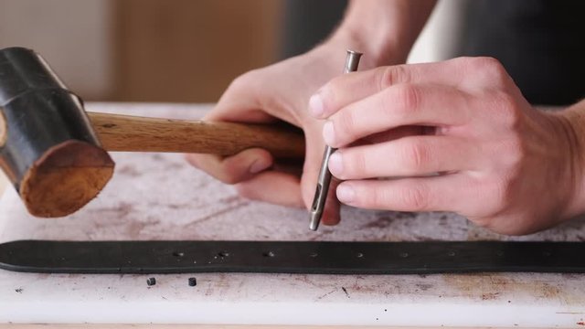 A leather craftsman is making holes in a hand-made belt. The tanner is using a mallet and a hole punch.
